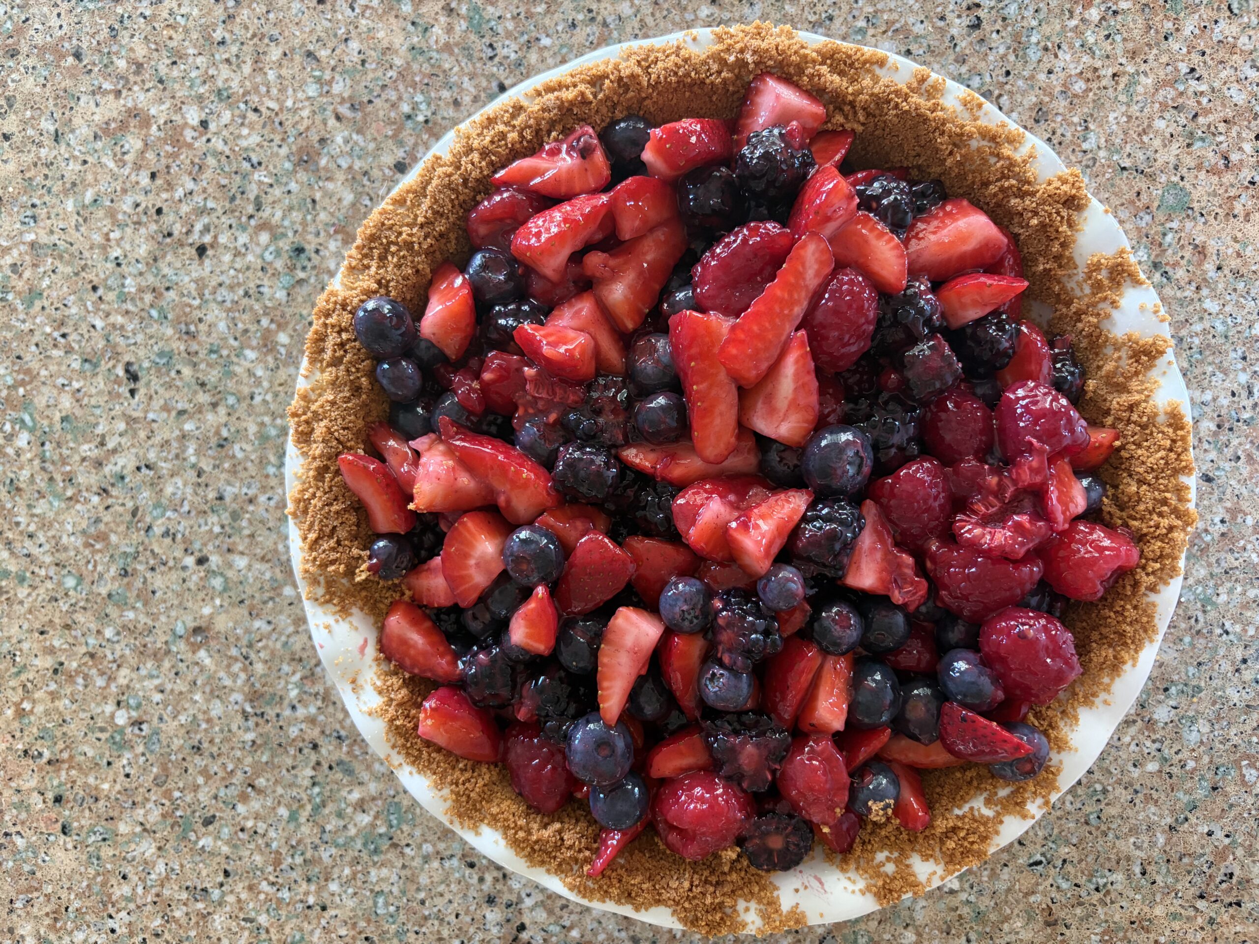 Mixed berry pie with strawberries, blackberries, blueberries, and raspberries in a graham cracker crust. Pie is sitting in a white pie plate on the counter.