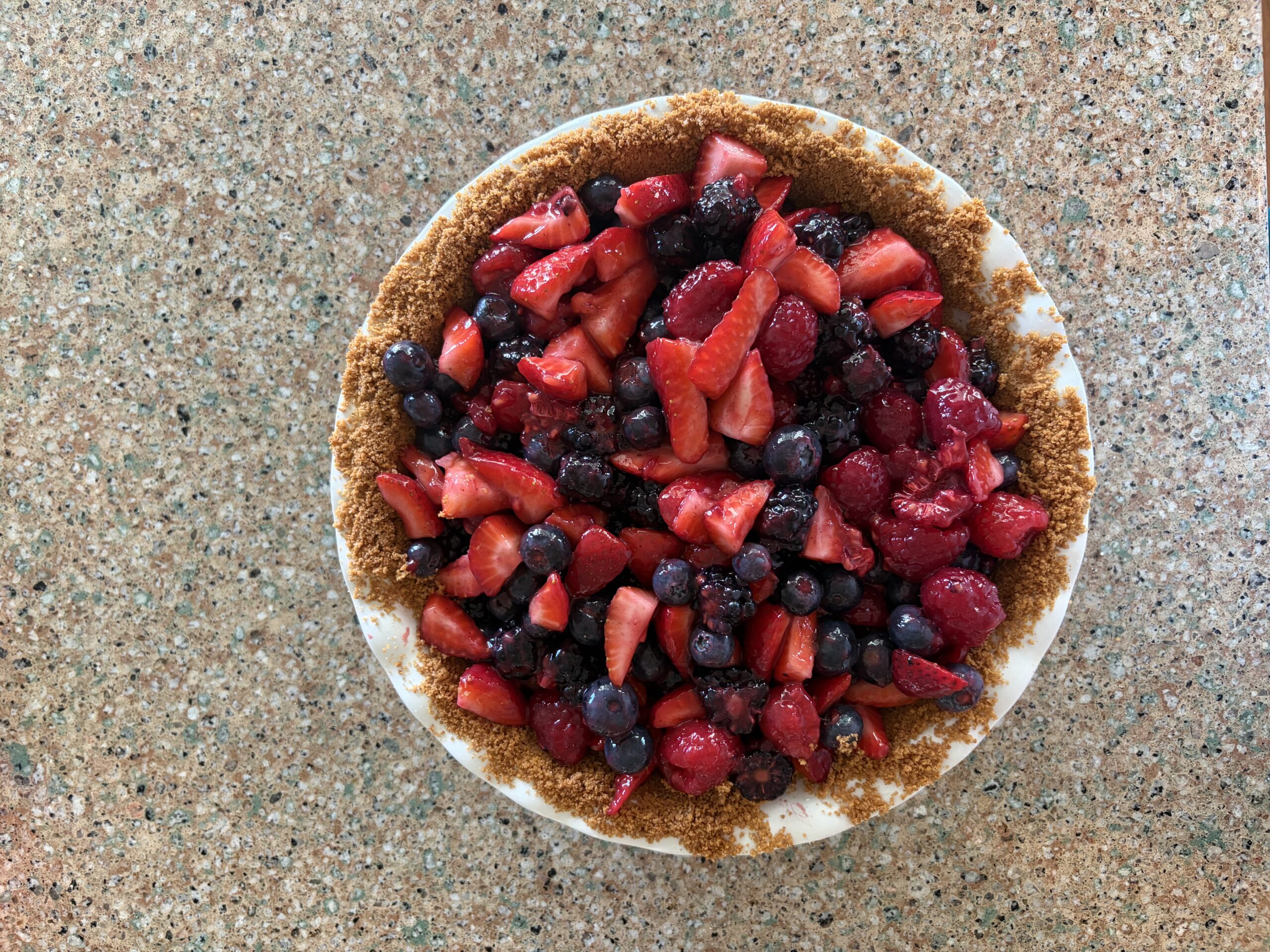 Mixed berry pie with strawberries, blackberries, blueberries, and raspberries in a graham cracker crust. Pie is sitting in a white pie plate on the counter.