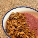 bowl filled with berry smoothie cover with granola that includes oats, puffed quinoa, 5 different types of nuts, and maple syrup.