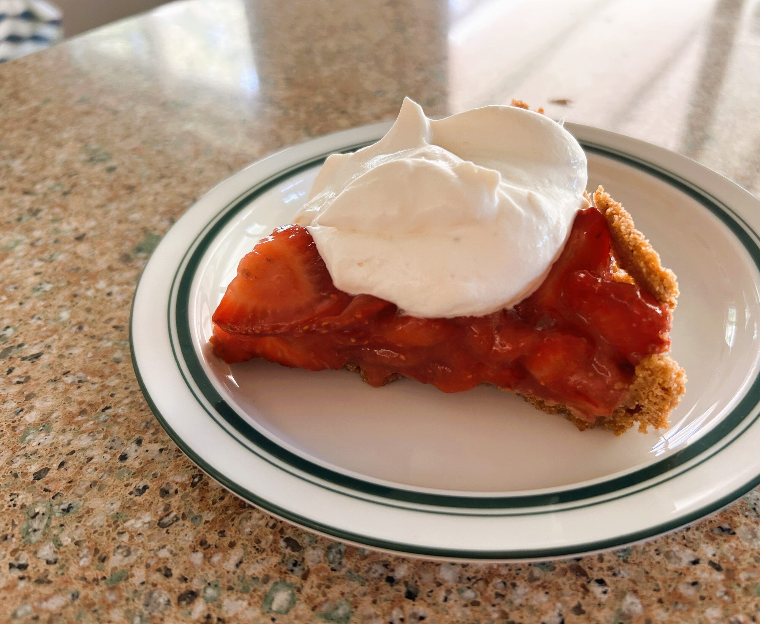 A slice of fresh strawberry pie with graham cracker crust, topped with fresh whipped cream on a plate sitting on a countertop.