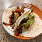 Two flour tacos sitting on a orange/white plate filled with roasted bell pepper, red onions, chicken, and black beans. Topped with sour cream and avocado.