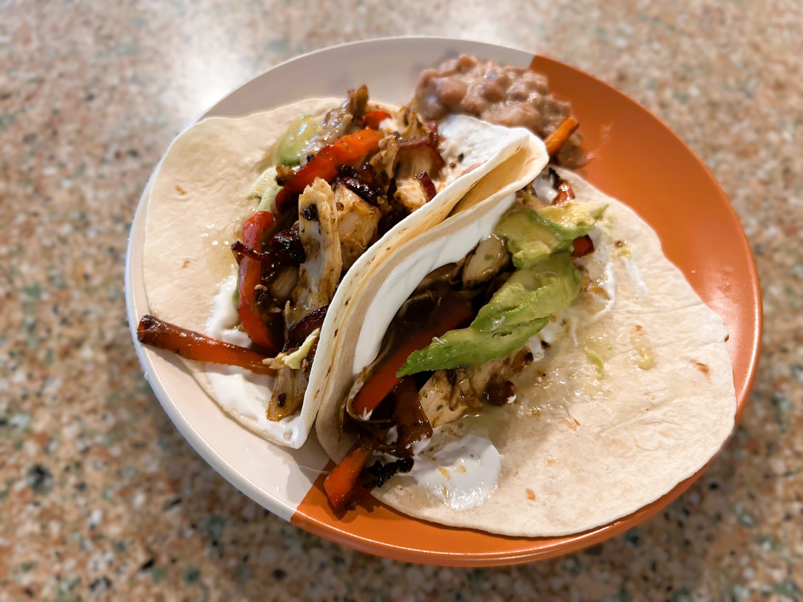 Two flour tacos sitting on a orange/white plate filled with roasted bell pepper, red onions, chicken, and black beans. Topped with sour cream and avocado.