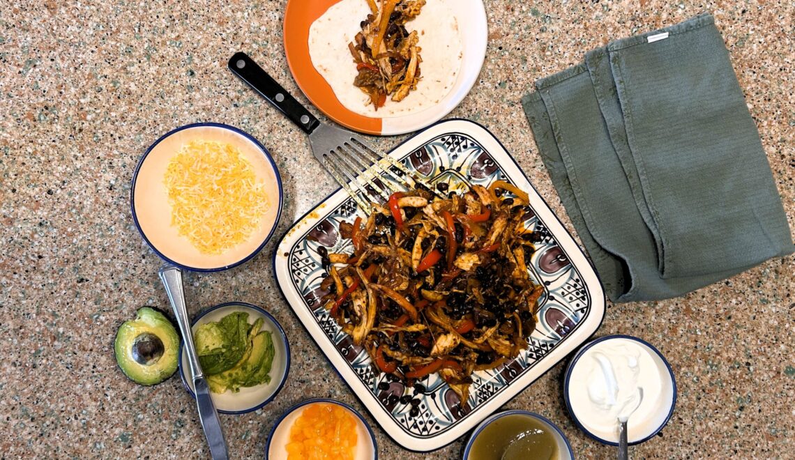 Fajitas on square plate, sour cream in a bowl, salsa in a bowl, cheese in a bowl, and avocado sliced in a bowl.