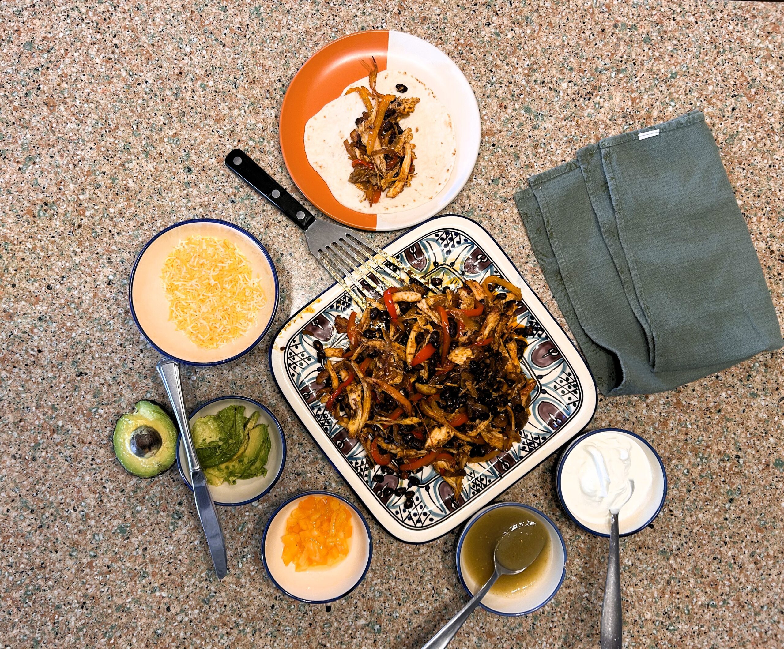 Fajitas on square plate, sour cream in a bowl, salsa in a bowl, cheese in a bowl, and avocado sliced in a bowl.