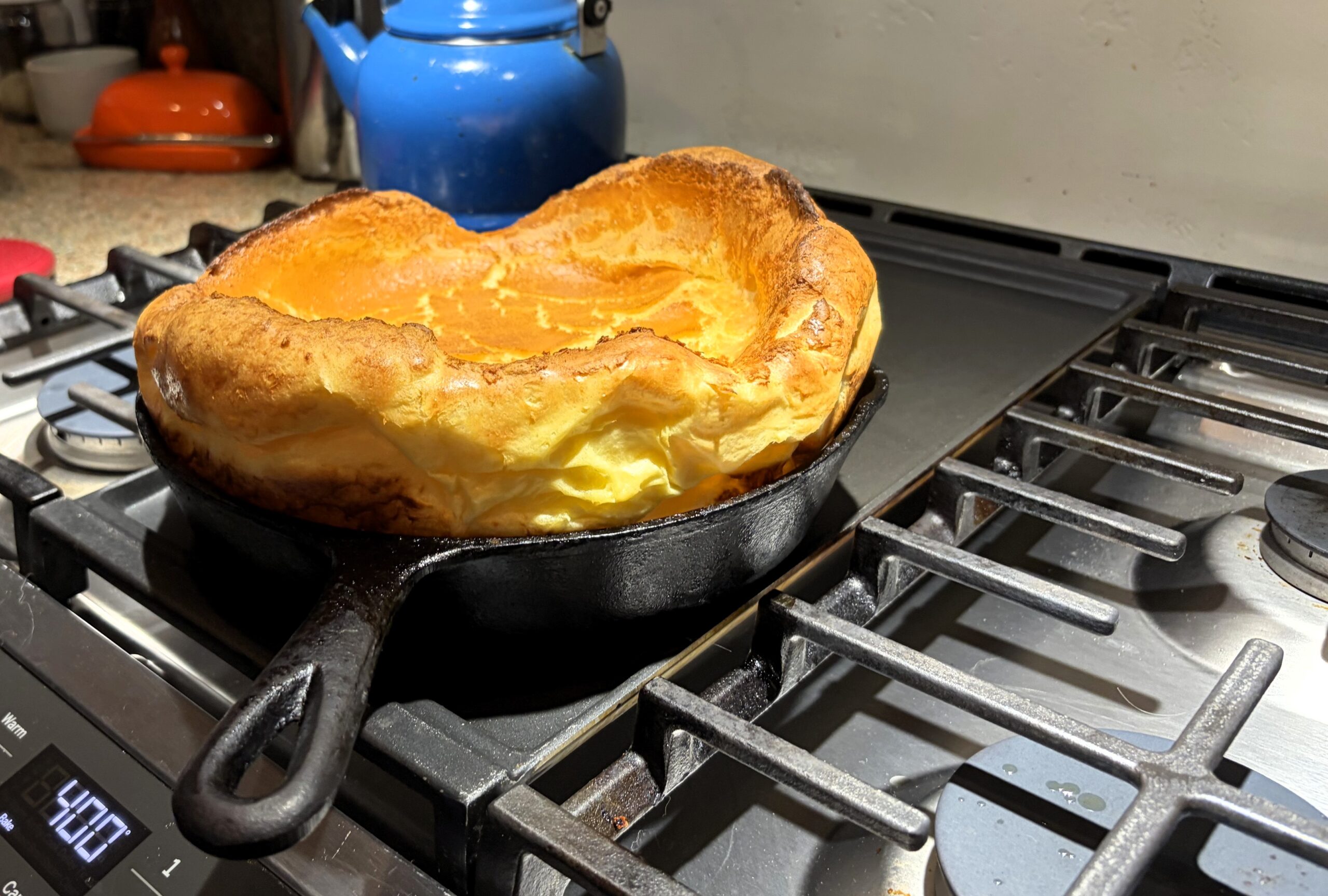 Baked dutch baby in the cast-iron pan sitting on the stove with a blue tea kettle in the background. 