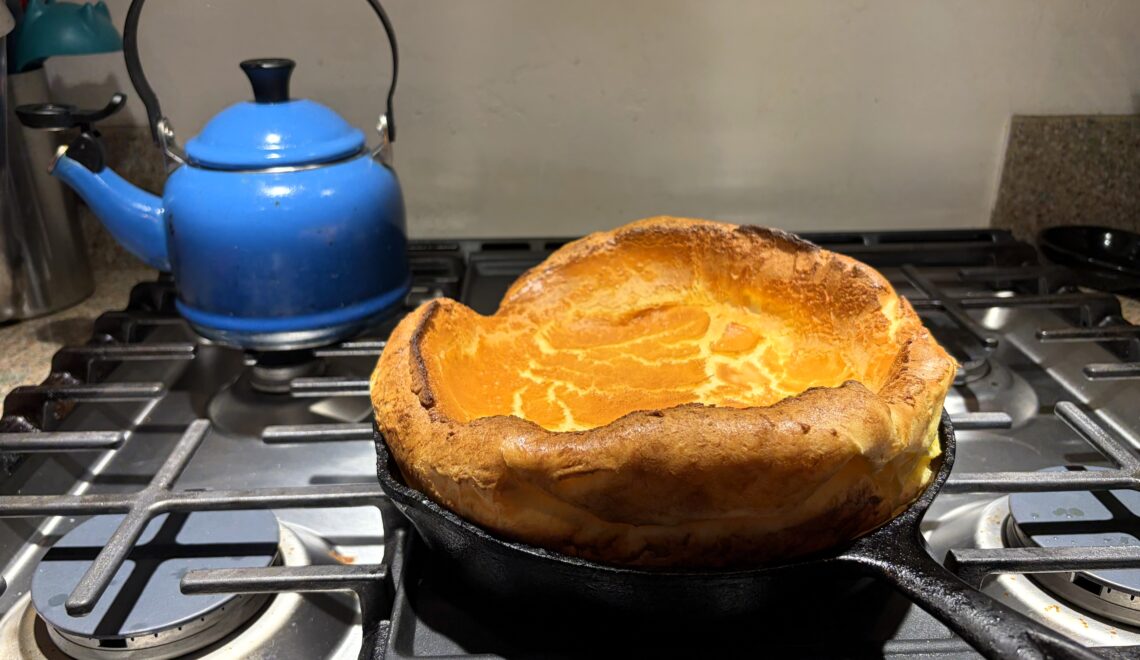 Dutch baby sitting on the stove in a cast-iron pan cooling, with a blue teakettle in the background.